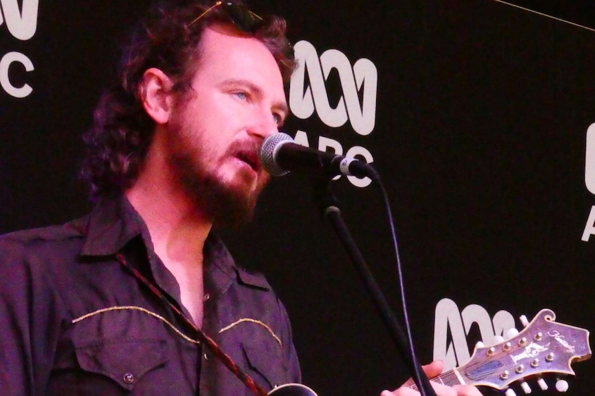 A man with long hair wears a black country shirt as he sings into a microphone and plays a mandolin on stage.