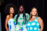 A woman of colour, a black woman and a white woman stand backstage at a fashion event wearing brightly-coloured dresses.