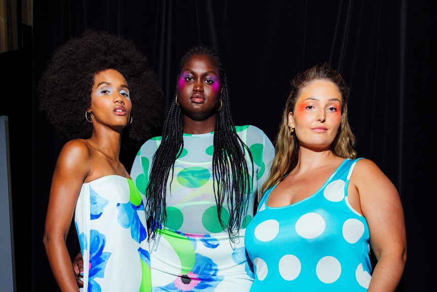 A woman of colour, a black woman and a white woman stand backstage at a fashion event wearing brightly-coloured dresses.