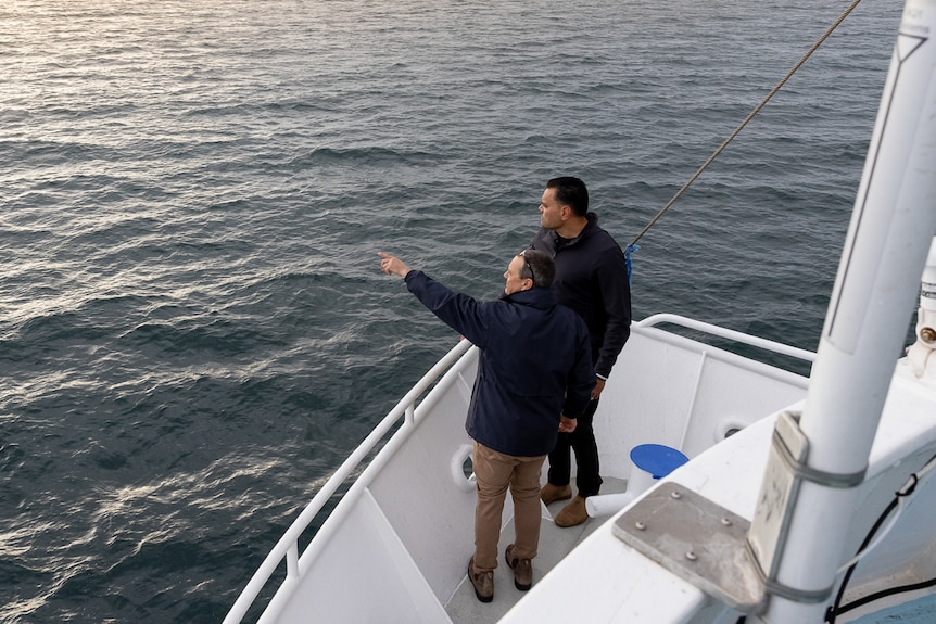 Two men stand at the front of the boat with one man pointing out to sea