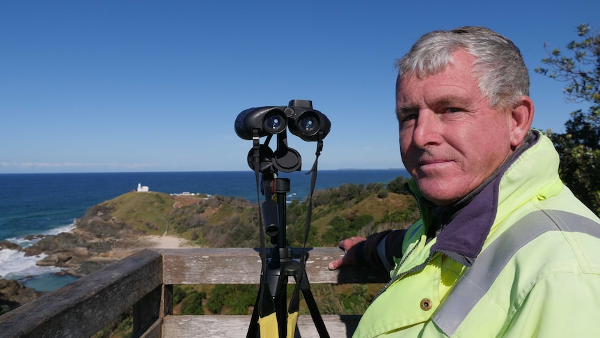 A man with grey hair and a bright yellow jacket stands on an ocean headland next to binoculars on a tripod.