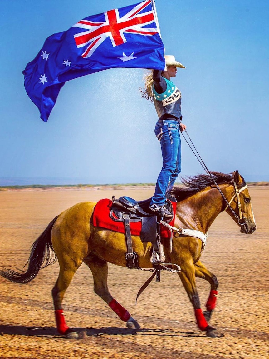 Woman stands on horseback holding up Australian flag