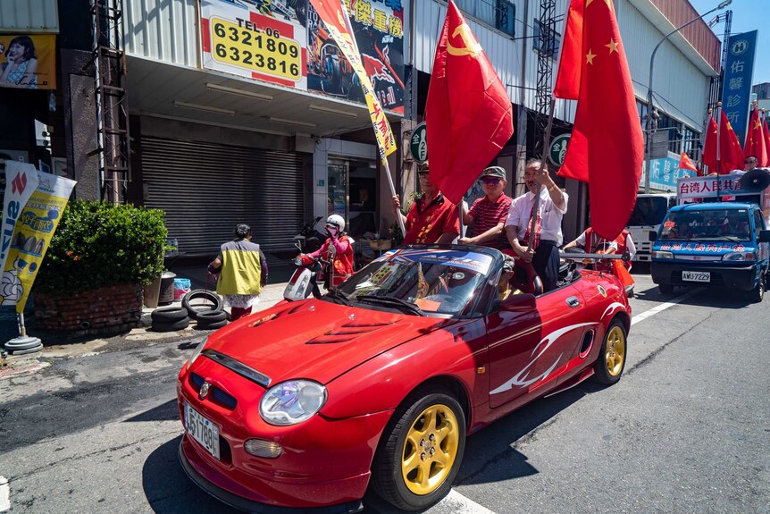 Three men wave red communists flags while standing in the back of a small red convertible car
