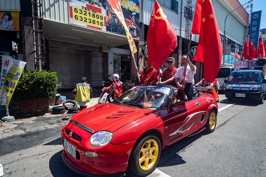 Three men wave red communists flags while standing in the back of a small red convertible car