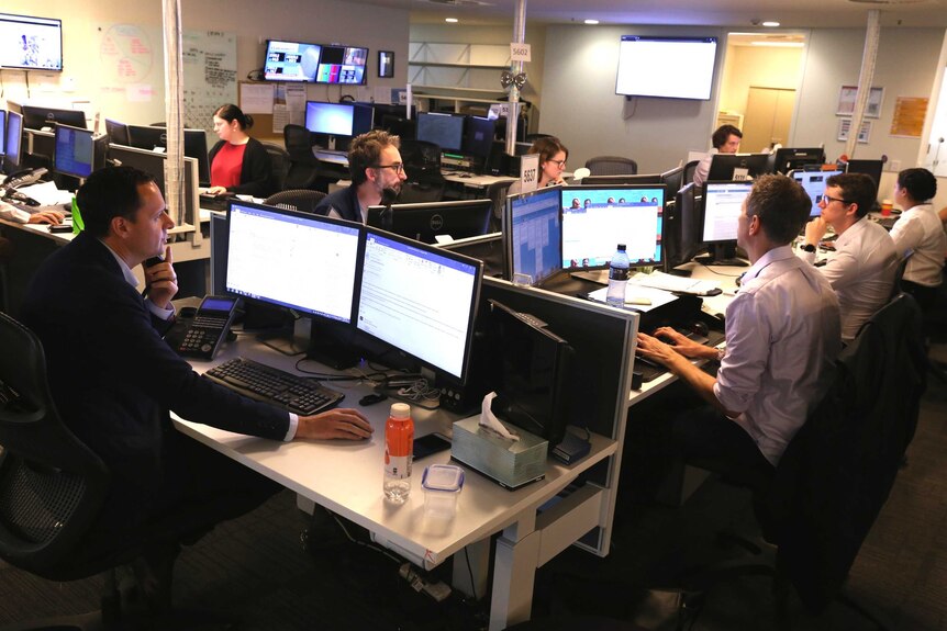 Wide shot of Sherlock at head of desk with numerous computer screens and members of team at work