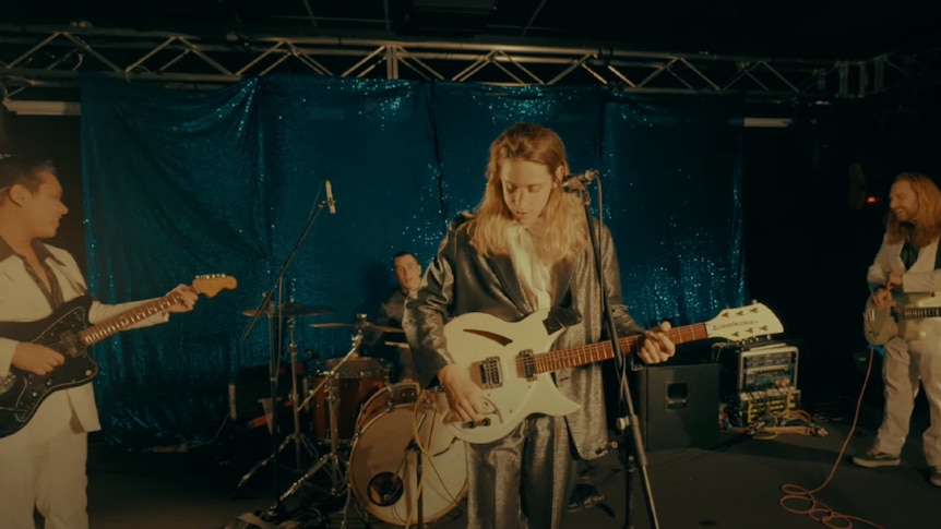 Three males dressed in beige suits, one in silver suit, holding guitars, bass and at a drum kit