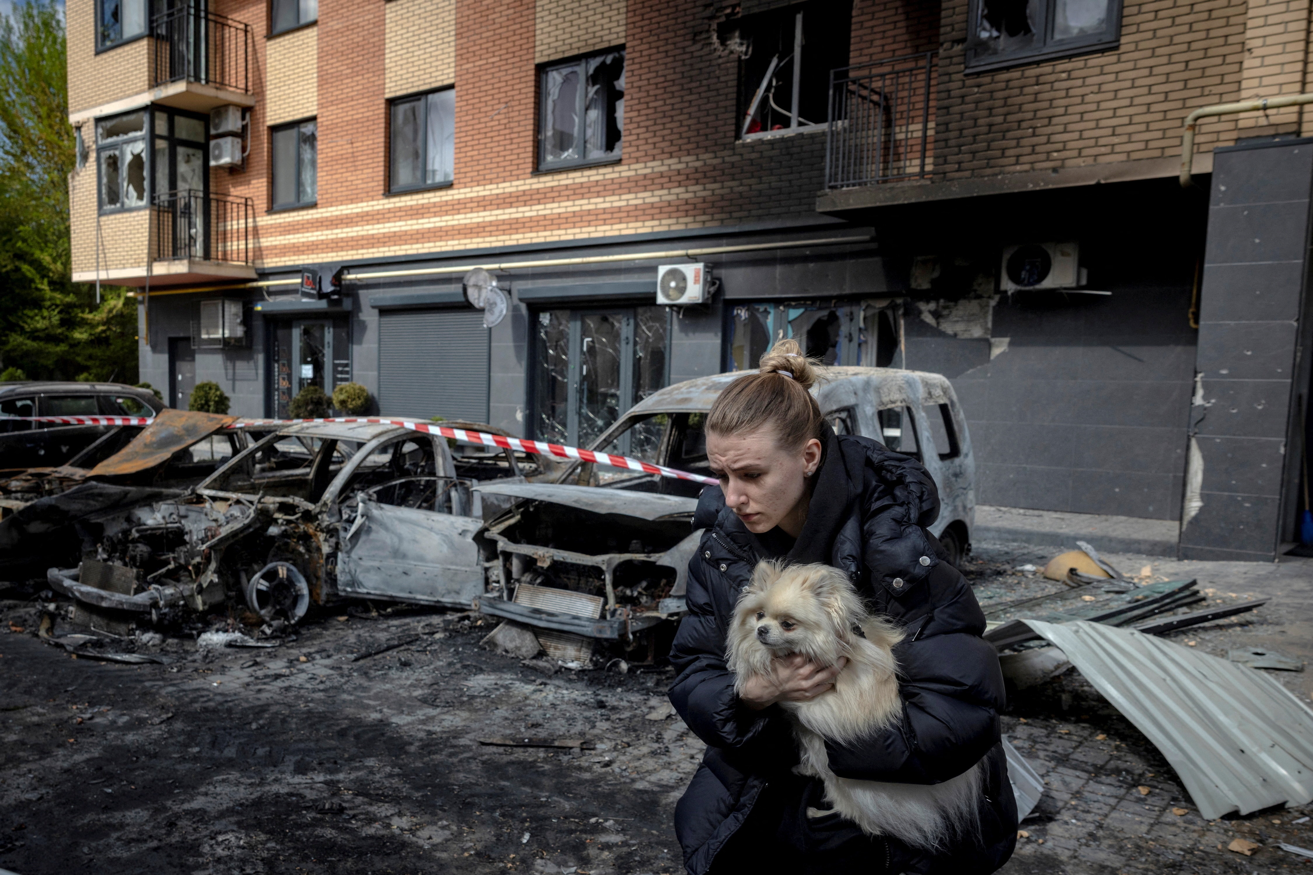 A woman and dog running out of a damaged apartment building with burnt-out vehicles parked out the front.