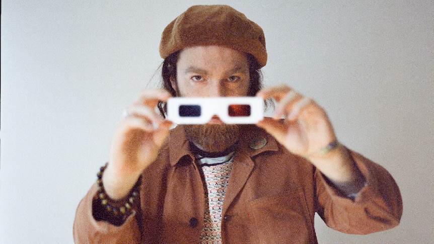 a man with beard holds 3D glasses, wearing brown beret and matching brown jacket