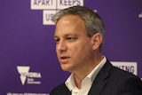 A man with short grey hair wearing a black suit jacket and white shirt stands in front of a purple backdrop.