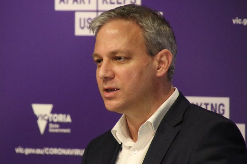 A man with short grey hair wearing a black suit jacket and white shirt stands in front of a purple backdrop.
