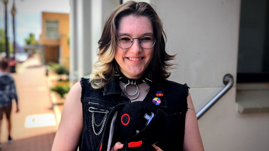 A white-skinned non-binary young person wearing glasses smiles at the camera, holding a plush toy.