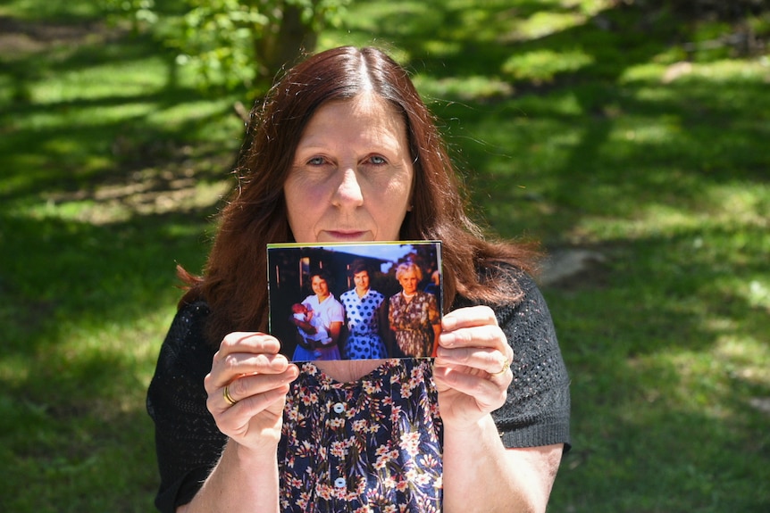 A woman with brown hair holds a photograph of three women, one holding a baby.