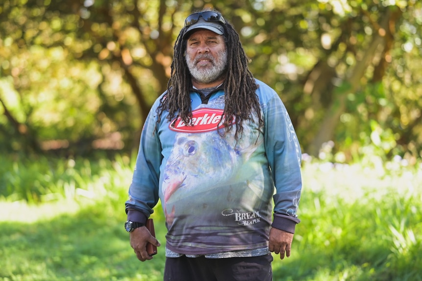 A man with dreadlocks and a cap stands in the bush.
