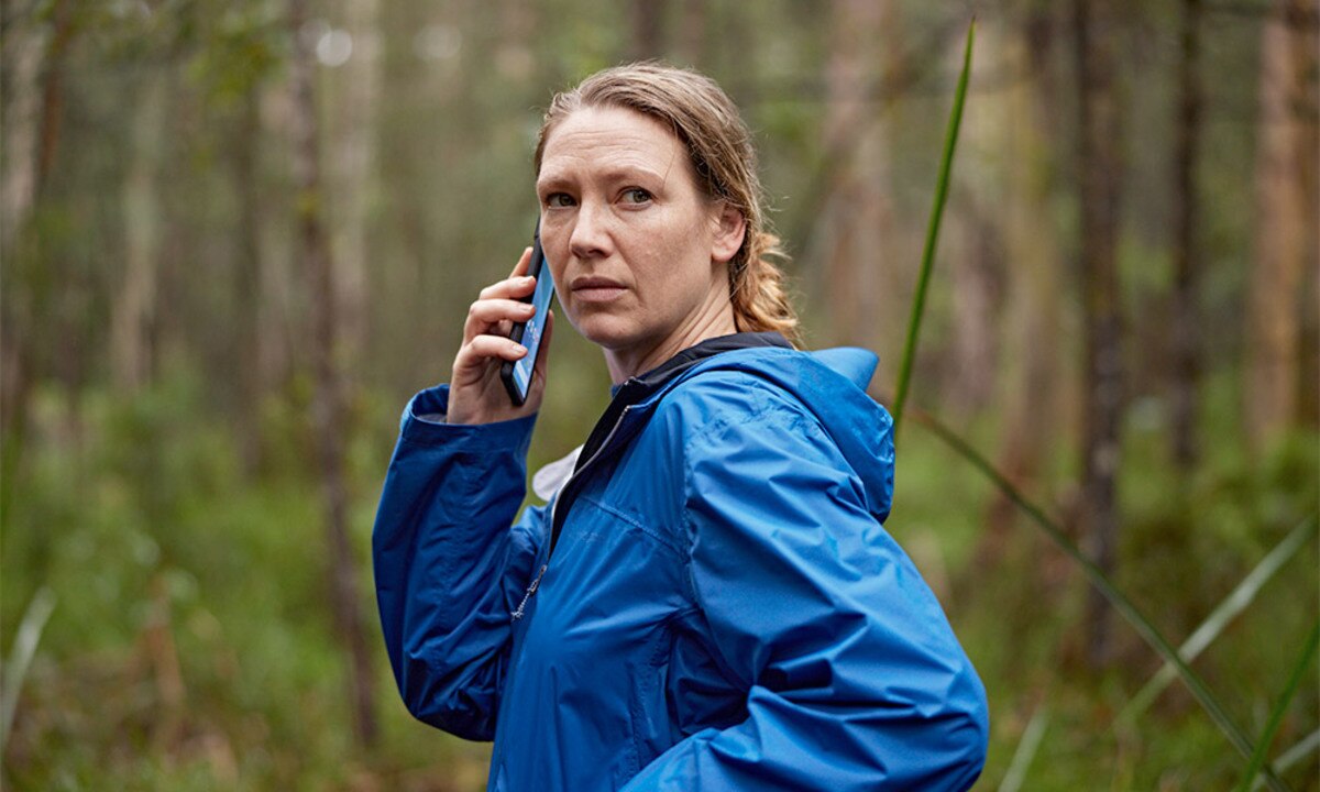 A woman in a blue raincoat stands with a phone to her ear, looking over her shoulder in the bush.