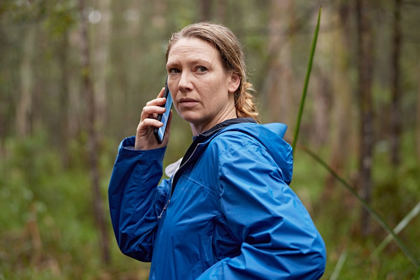 A woman in a blue raincoat stands with a phone to her ear, looking over her shoulder in the bush.