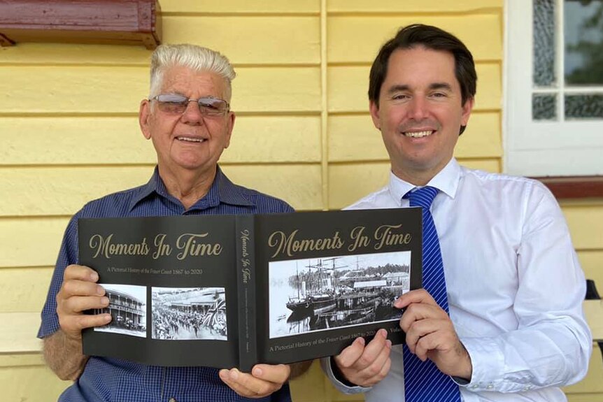 Two men sit looking at the camera smiling and holding a book about the history of the Fraser Coast.