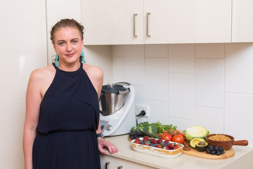 A woman stands in her kitchen besides a benchtop covered in fresh food.