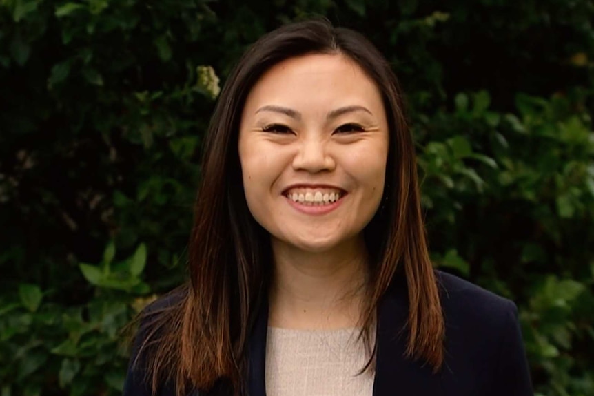 A woman in a cream shirt and dark blue blazer with long straight brown hair speaks with green bushes in the background.