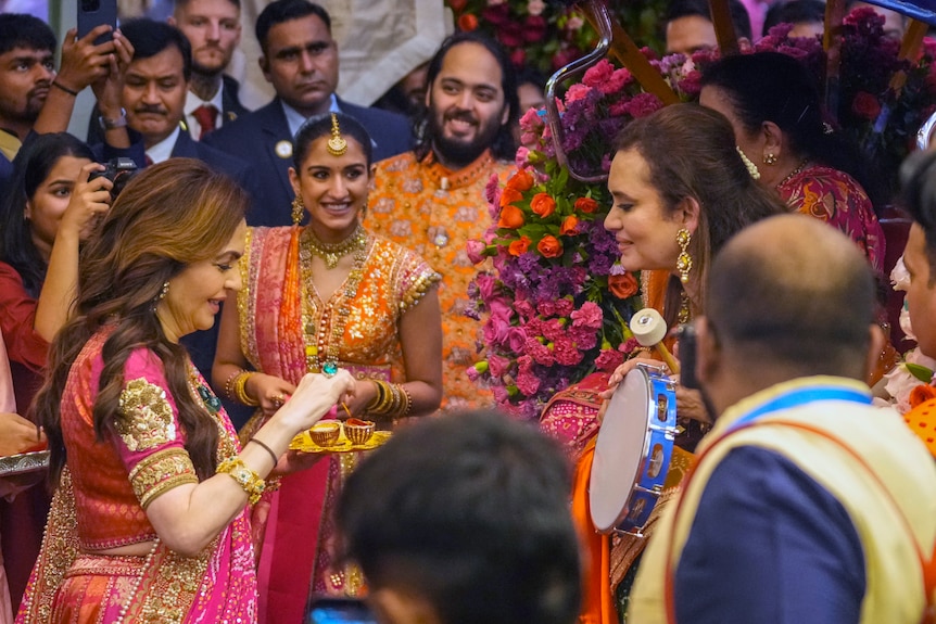 Nita Ambani greets a guest with an offering as well-dressed onlookers stand by.