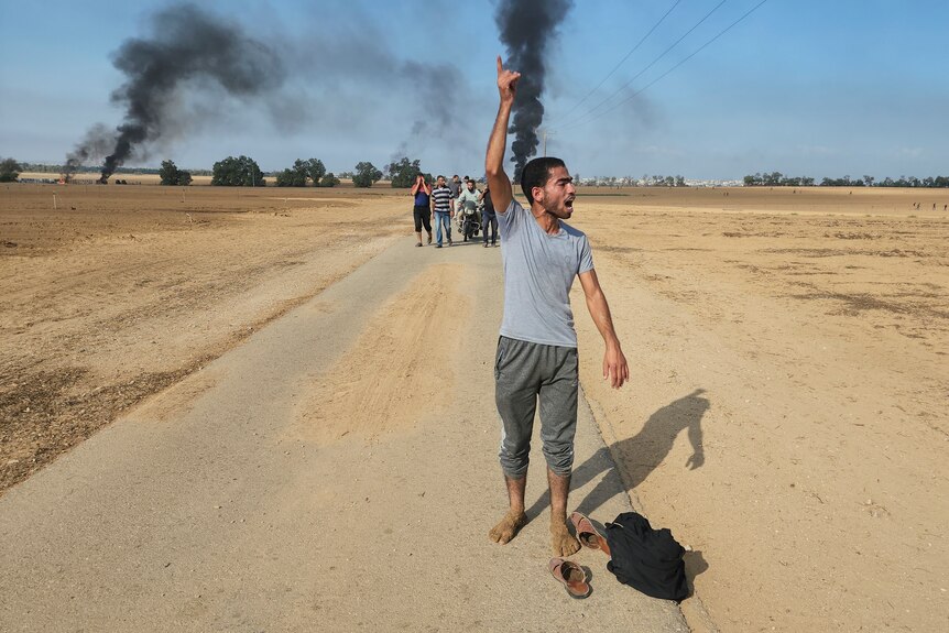 A man walks along a road with his arm in the air as a group of people walk behind him, and behind him smoke rises