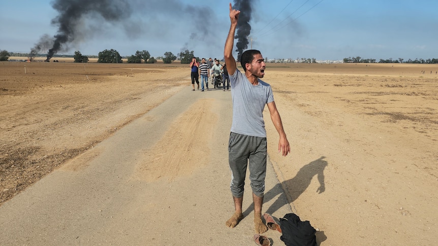 A man walks along a road with his arm in the air as a group of people walk behind him, and behind him smoke rises