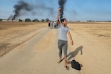 A man walks along a road with his arm in the air as a group of people walk behind him, and behind him smoke rises