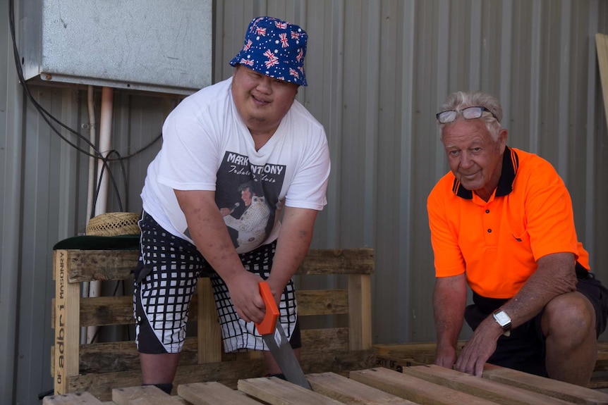 A man bending over holding a saw cutting into a wooden pallet while another man kneels beside wearing high visibility gear