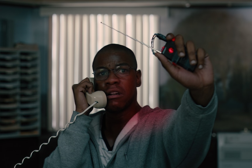 Movie still: Man in hoodie holds phone to his ear and holds up explosive remote.