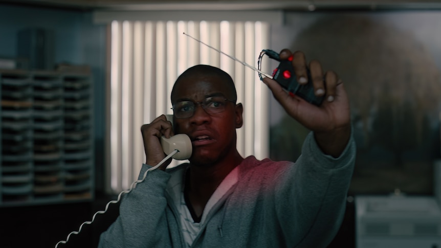Movie still: Man in hoodie holds phone to his ear and holds up explosive remote.