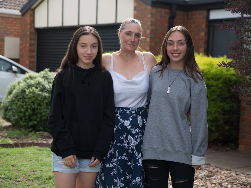 Linda has her arms around her two daughters as they stand in front of their brown brick home