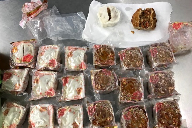 Twenty mooncakes - pastry on outside filled with pork - on a bench. Some are wrapped in plastic. Two are opened.
