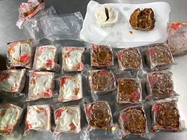 Twenty mooncakes - pastry on outside filled with pork - on a bench. Some are wrapped in plastic. Two are opened.