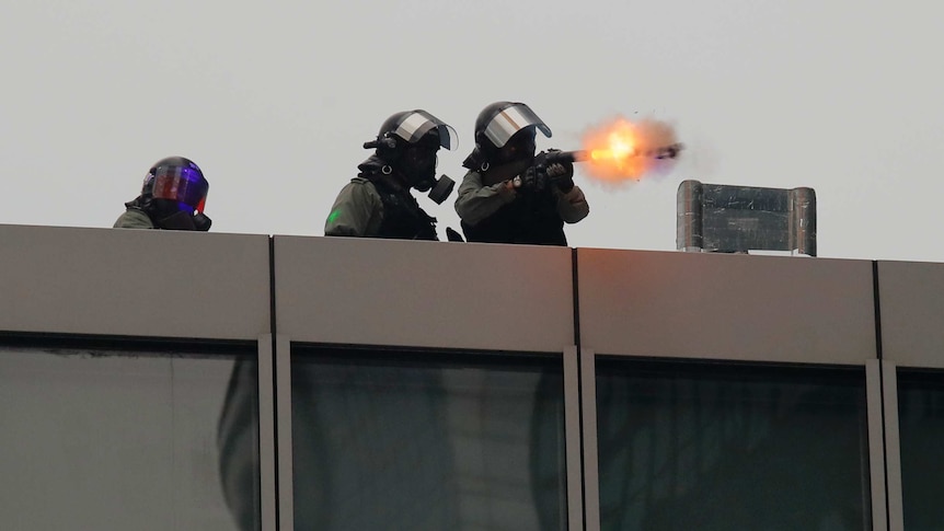 flames are seen coming from the end of a gun fired by police wearing riot gear