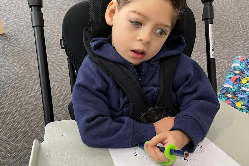 Little boy in harness chair holding pen and doing drawing at table