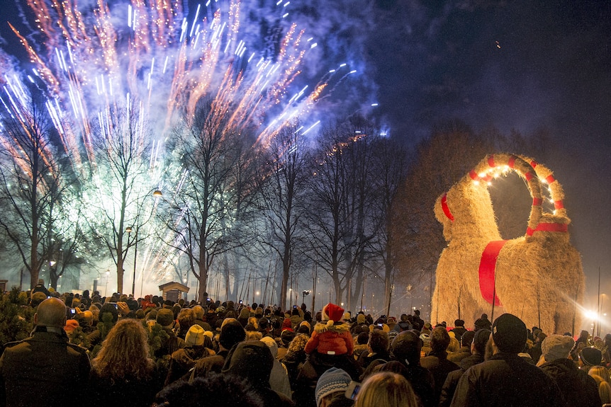 People gather outside in a public square at the inauguration of a giant straw goat while watching colourful fireworks.