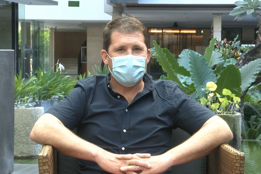 A man in a blue face mask and black shirt sits in a tropical garden 