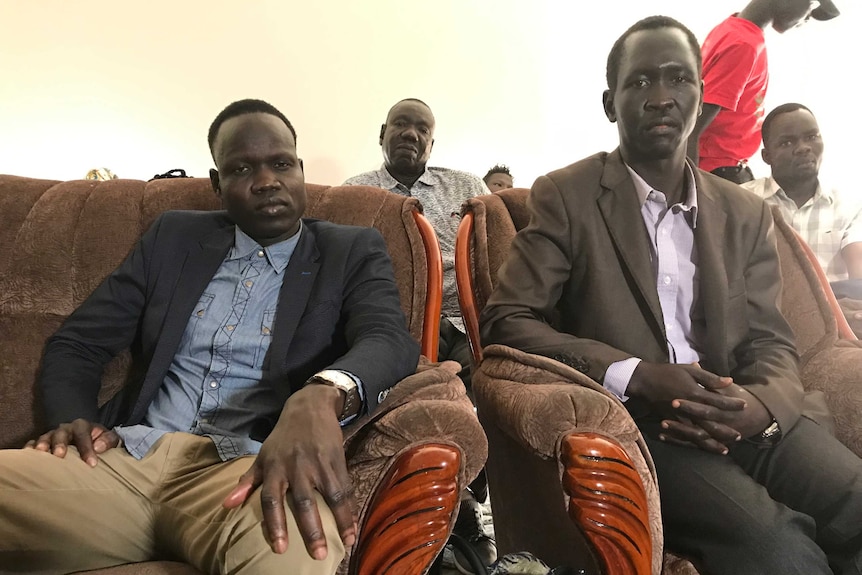 Laul Akec and Peter Riak sit on brown armchairs looking at the camera with sad expressions with family in the background.