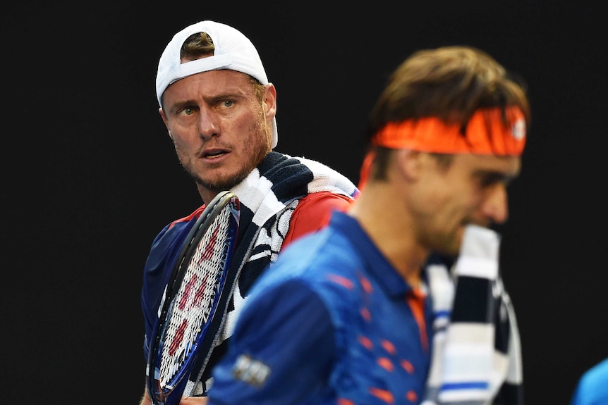 Lleyton Hewitt is in focus as David Ferrer walks in front of him, blurred in the foreground.
