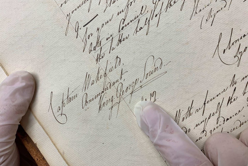 Cursive writing on an old ledger, held by a person wearing gloves and pointing to the name King George Sound.