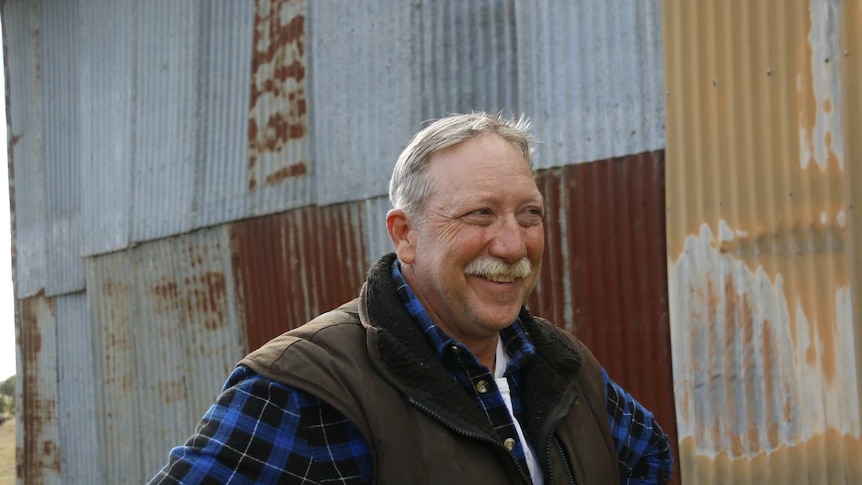 Man with grew hair and a grey moustache smiles against a rusty shed backdrop.