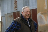Man with grew hair and a grey moustache smiles against a rusty shed backdrop.