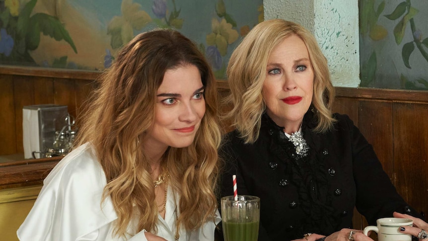 Alexis and Moira sitting at a table in the cafe in the TV show Schitt's Creek in a story about parenting lessons from Moira.