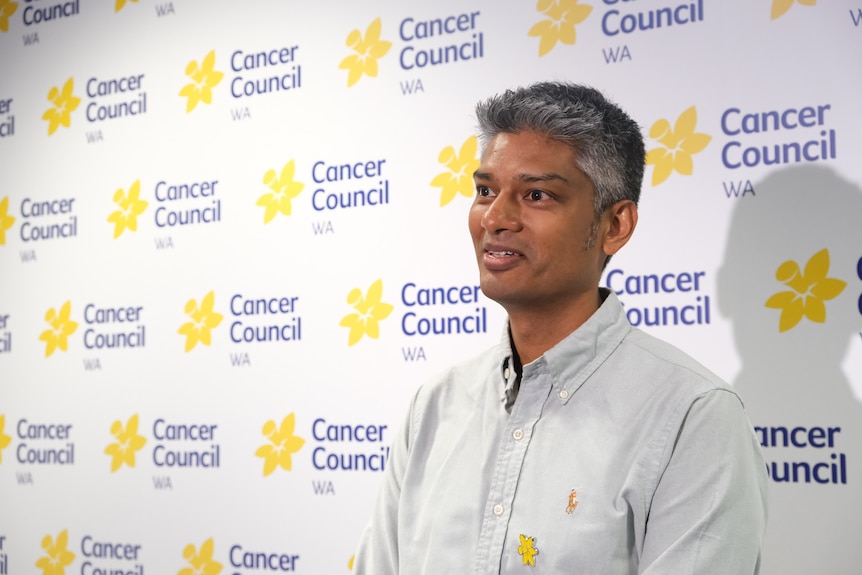A man with dark skin and a light collared shirt stands against a Cancer Council WA backdrop
