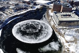 A view from above of a large ice disk floating in a river in a wintery urban landscape