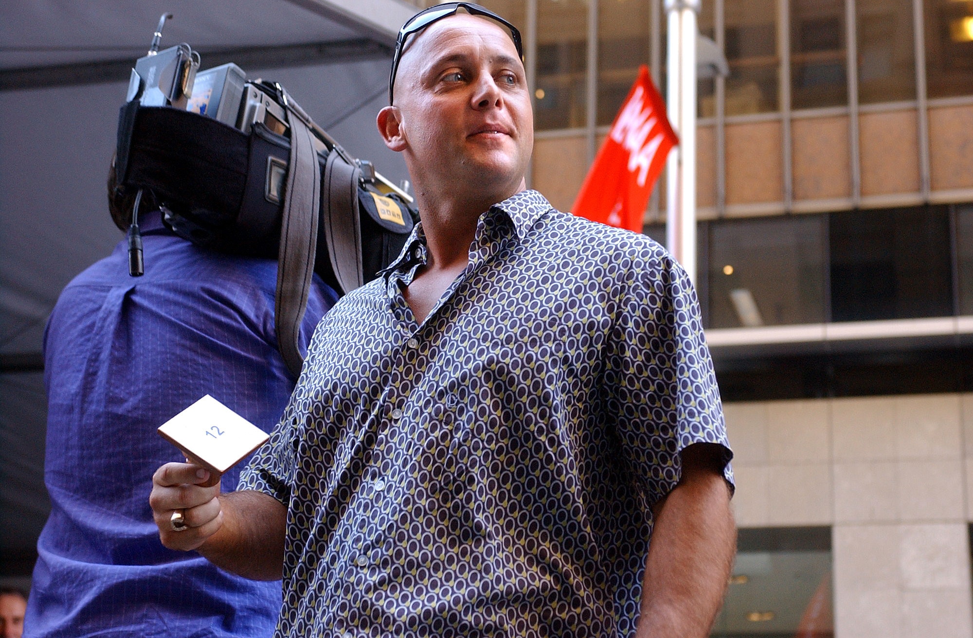 A bald man in a short-sleeved shirt holds a card and looks to the side.