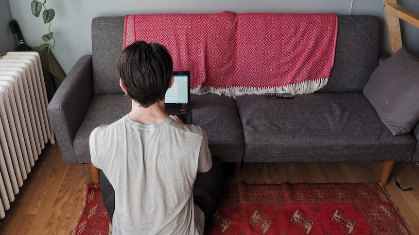 A man sits on the floor, his back to the camera, typing on his laptop which is perched on a couch