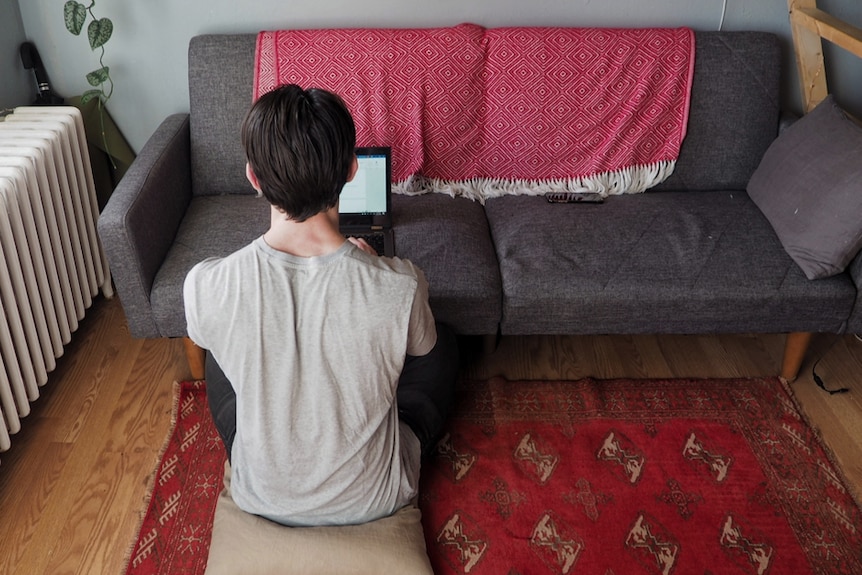 A man sits on the floor, his back to the camera, typing on his laptop which is perched on a couch