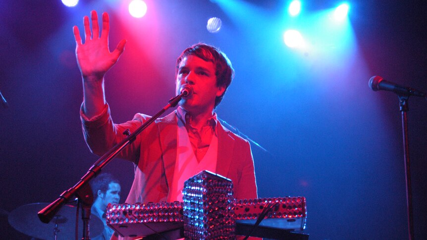 Brandon Flowers stands behind a jewell encrusted keyboard, his arm outstretched and palm open