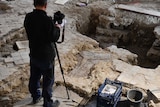 A man standing on a stone floor with a camera tripod. Under his feet is a coffin-shaped rock.