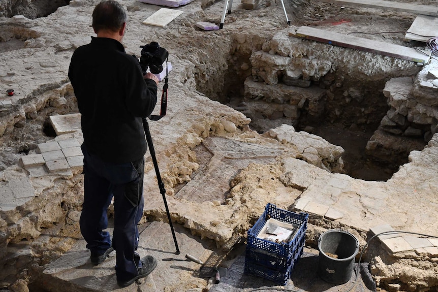 A man standing on a stone floor with a camera tripod. Under his feet is a coffin-shaped rock.